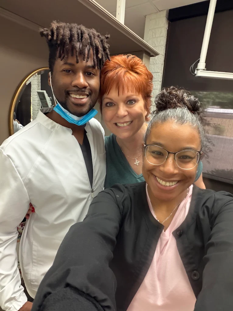 Three smiling people posing for a selfie in a hair salon setting with shelves behind them.