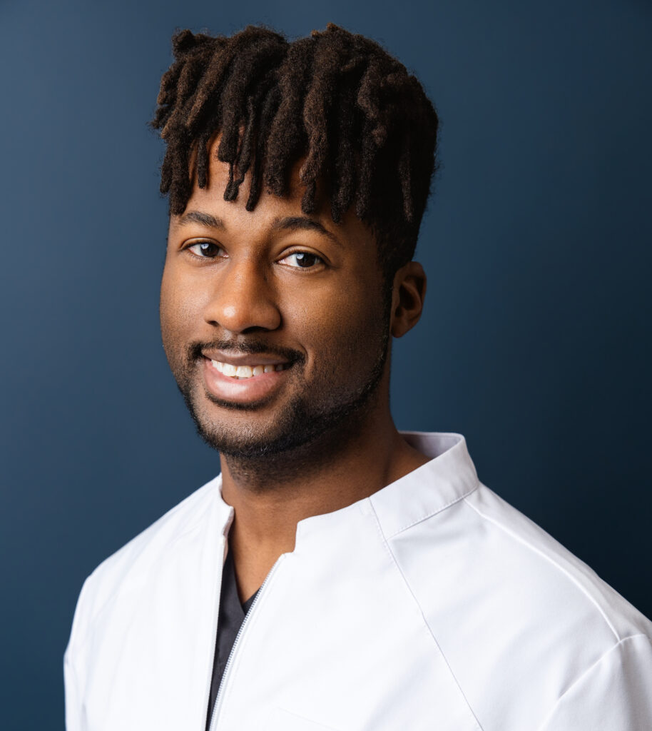 Close-up of a smiling man with short dreadlocks, wearing a white coat against a blue background.